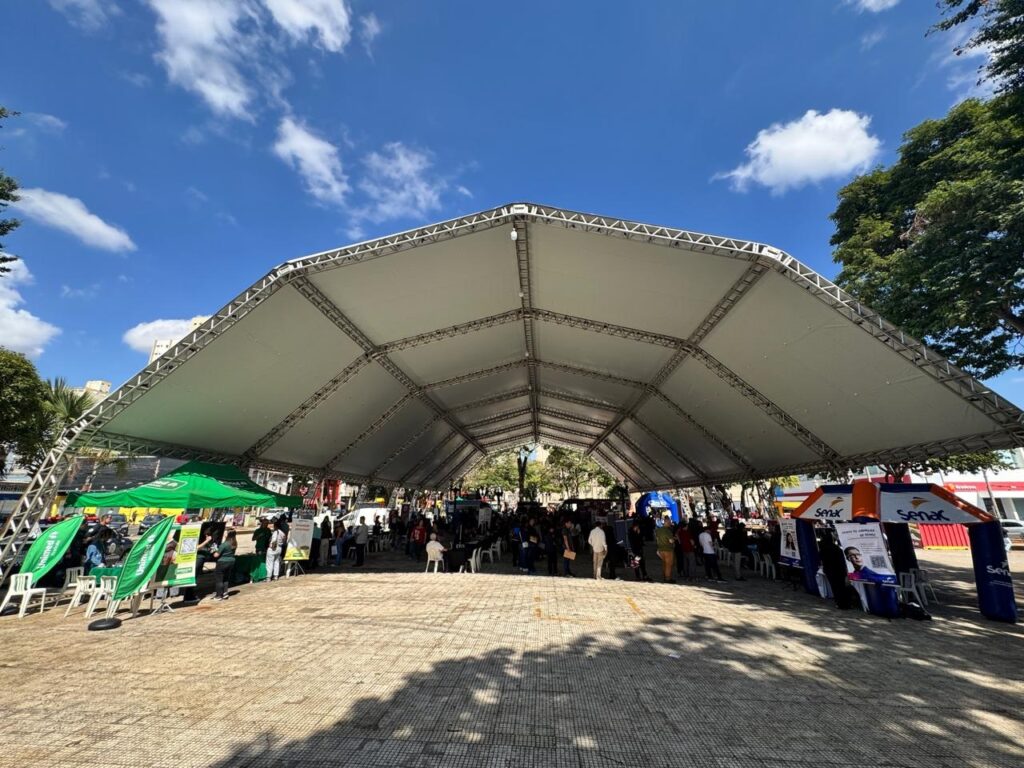 Esta imagem mostra uma feira, evento ou ação de serviços ao ar livre, montada em uma praça pública sob uma grande tenda estruturada. Aqui estão os detalhes principais: Estrutura: Há uma tenda branca grande e ampla cobrindo uma área de piso de pedras claras (pavimentação de praça), proporcionando sombra para os participantes. Expositores: Vários estandes e tendas menores estão montados sob a cobertura principal. É possível identificar marcas como Unimed (à esquerda, com tenda verde) e Senac (à direita, com estrutura azul e laranja). Público: Várias pessoas estão circulando pelo local, conversando nos estandes e caminhando pelo espaço, indicando um dia de atendimento ao público. Ambiente: O céu está azul e ensolarado, com algumas nuvens, e há árvores nas laterais da praça, sugerindo um clima agradável. O ambiente parece ser urbano, com prédios ao fundo e uma atmosfera de evento organizado durante o dia.