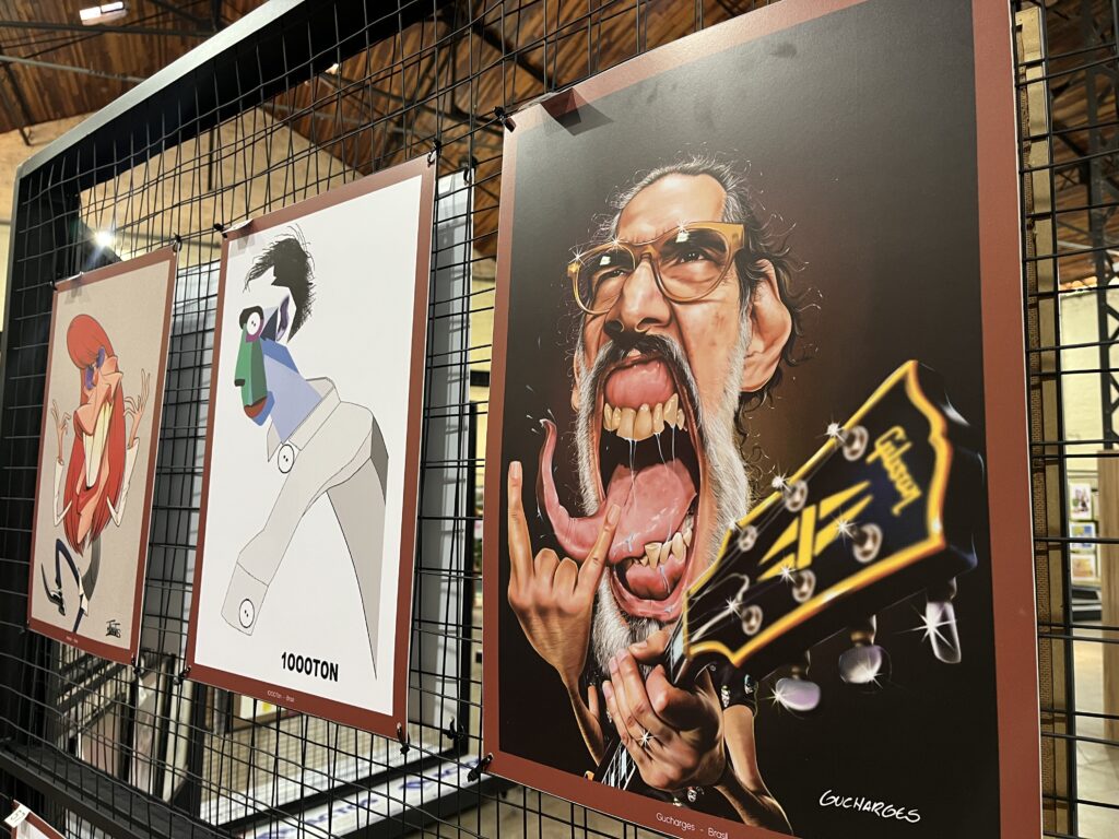 Na imagem, aparecem três obras em estilo caricatural expostas em uma grade metálica preta, como parte de uma mostra de arte. Cada impressão apresenta retratos exagerados e criativos: à esquerda, uma figura de cabelos vermelhos longos e corpo alongado em pose dinâmica; ao centro, um rosto geométrico e abstrato com traços angulares e cores vibrantes, acompanhado da inscrição “1000TON”; e à direita, um personagem com óculos, bigode e boca aberta em grito, segurando uma guitarra Gibson, assinado “GUCHARGES”.