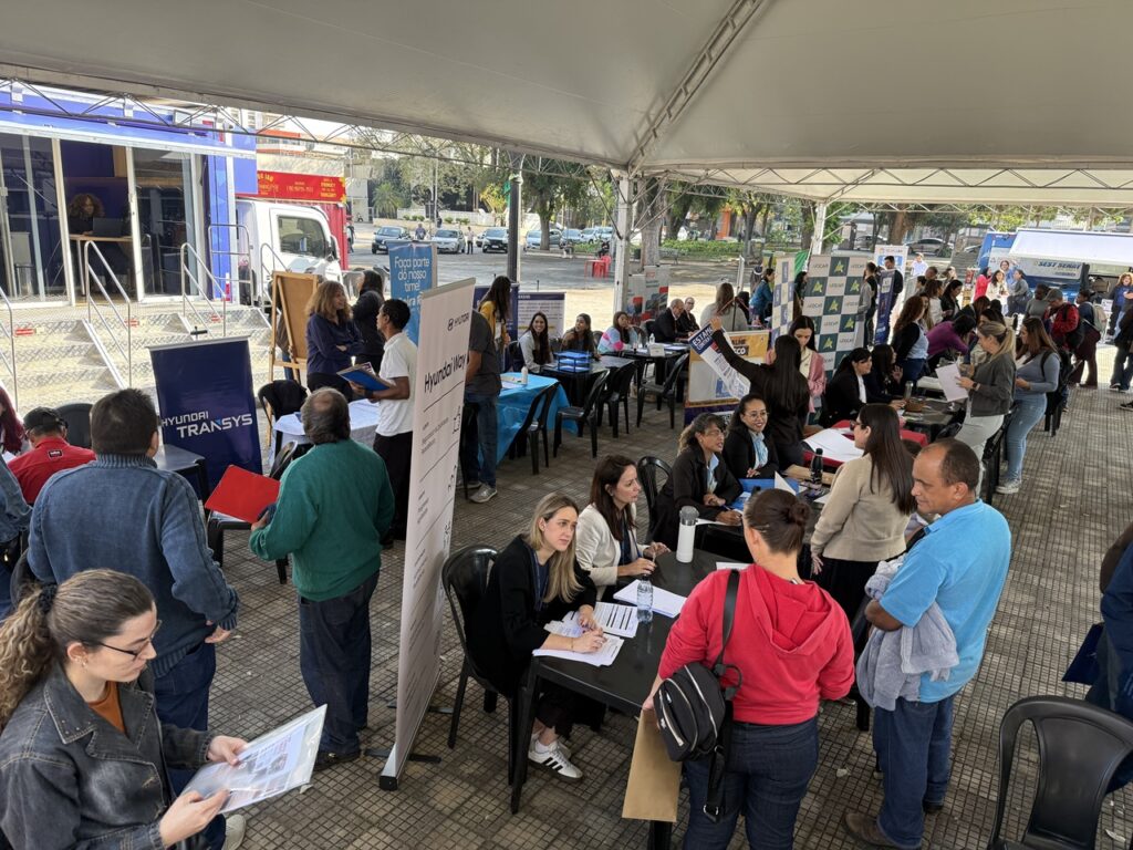 A imagem mostra um evento movimentado sob uma grande tenda branca, com várias mesas organizadas em fileiras e pessoas conversando em um ambiente de feira de empregos. Participantes seguram currículos e formulários enquanto representantes de empresas e instituições — como Hyundai Transys, Hyundai Motor e ACIA — atendem interessados.