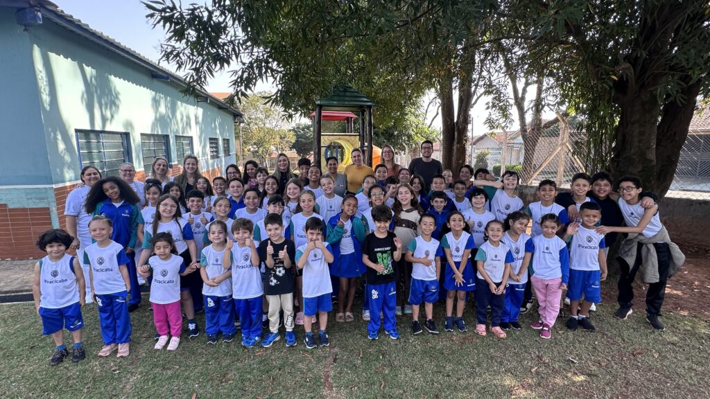 vemos um grande grupo de crianças e adultos reunidos em frente a uma estrutura de playground e ao lado de um prédio de paredes azuis. As crianças estão uniformizadas com camisetas brancas de detalhes azuis ou verdes e calças azuis, muitas trazendo a inscrição “Piracicaba”, o que sugere tratar-se de uma turma escolar. Atrás delas, os adultos — provavelmente professores ou funcionários — acompanham a cena. O ambiente é um pátio escolar, com grama, árvores que oferecem sombra e um cercado ao fundo.