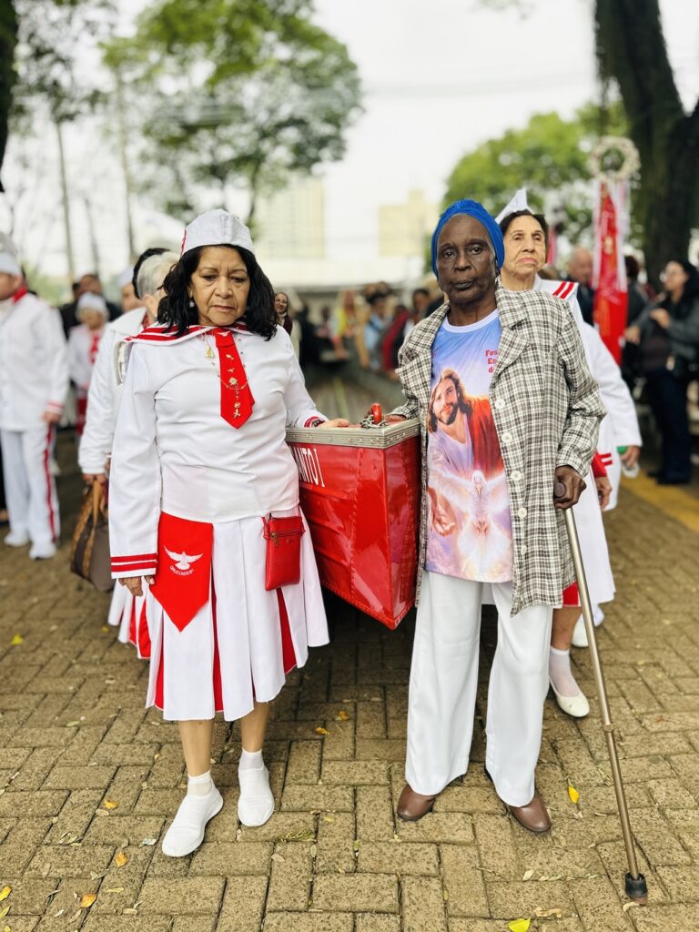 Na imagem há um grupo de pessoas participando de uma procissão ou evento religioso/cerimonial ao ar livre. Em primeiro plano, duas mulheres caminham lado a lado: À esquerda: mulher com traje branco e detalhes vermelhos (saia plissada, gravata/lenço vermelho, pequeno bolso/bolsa vermelha), usando também um chapéu branco. Ela segura a alça de uma caixa vermelha grande — a caixa tem partes metálicas e parece ser usada para transporte de objetos do grupo. À direita: mulher negra, de idade avançada, usando lenço azul na cabeça, casaco xadrez claro por cima de uma camiseta com imagem religiosa (figura de Jesus) e calça branca. Apoia-se em uma bengala metálica com ponteira de borracha. Ao fundo e nas laterais: outras pessoas vestindo uniformes brancos com faixas/vermelhos semelhantes, algumas com chapéus; há estandartes e bandeiras vermelhas, sugerindo organização e identidade comum (grupo religioso ou filantrópico). Ambiente: passeio de paralelepípedo com árvores e pessoas formando uma fila; clima ameno/iluminado por luz natural difusa, provavelmente manhã ou tarde nublada. Expressões e postura: as duas mulheres em primeiro plano têm expressões sérias ou concentradas; a cena transmite solenidade, respeito e participação comunitária. Detalhes visuais: folhas caídas no chão, prédios desfocados ao fundo e outras pessoas observando/participando, compondo um cenário de evento público.