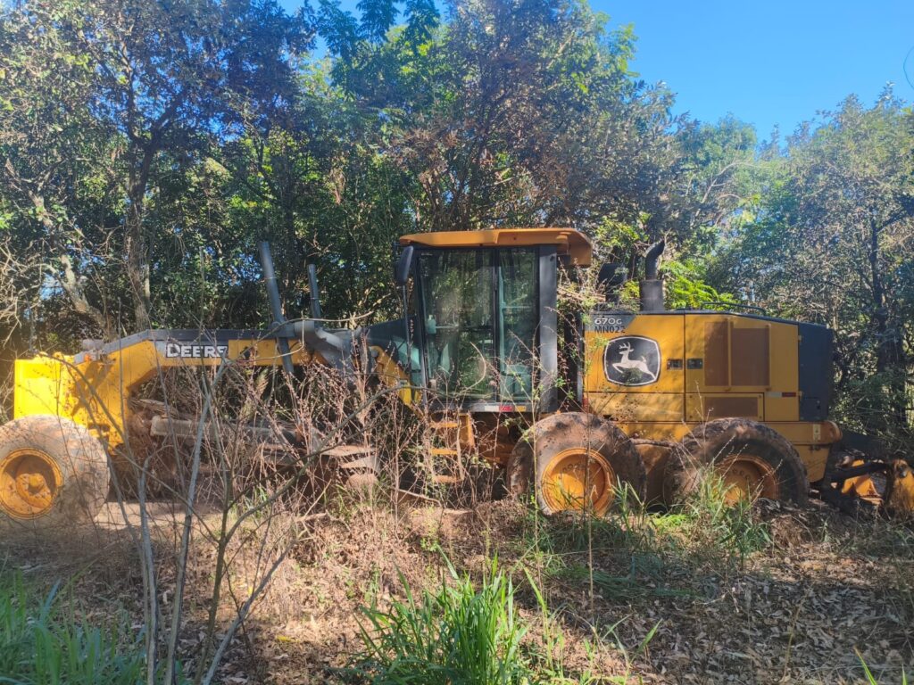 Nesta imagem vemos uma motoniveladora John Deere, modelo 670G, parada em uma área de vegetação, possivelmente na beira de uma estrada de terra ou canteiro de obras. Aqui estão os detalhes principais: Equipamento: É uma motoniveladora amarela, típica de obras de terraplanagem e manutenção de estradas, com o logotipo característico do cervo da marca John Deere visível. Estado: O equipamento parece estar em uso ou operando recentemente, já que há terra acumulada nos pneus e na estrutura. Ambiente: Ela está posicionada entre árvores e arbustos, o que sugere um contexto rural ou de trabalho em áreas de abertura de via. Identificação: É possível ver claramente o modelo "670G" e uma inscrição "MN022" na lateral.