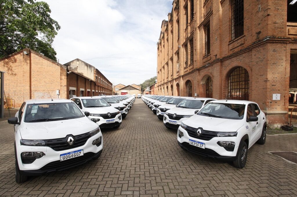 Esta imagem registra uma frota numerosa de carros brancos, modelo Renault Kwid, estacionados de forma perfeitamente alinhada em duas fileiras paralelas, em um pátio pavimentado com blocos de concreto. O cenário: A frota ocupa o espaço central entre dois grandes prédios de tijolos aparentes, com arquitetura que lembra antigas fábricas ou armazéns, caracterizados por arcos nas janelas e uma estrutura robusta e histórica. A organização: O alinhamento dos veículos, todos virados na mesma direção, cria um efeito visual de simetria e organização, sugerindo uma entrega oficial, inspeção ou renovação de frota de uma instituição, como a Prefeitura de Piracicaba (identificável pelos logotipos e numerações nas laterais dos carros). O ambiente: O dia parece claro, com céu parcialmente nublado, e o espaço transmite uma sensação de escala e importância institucional.