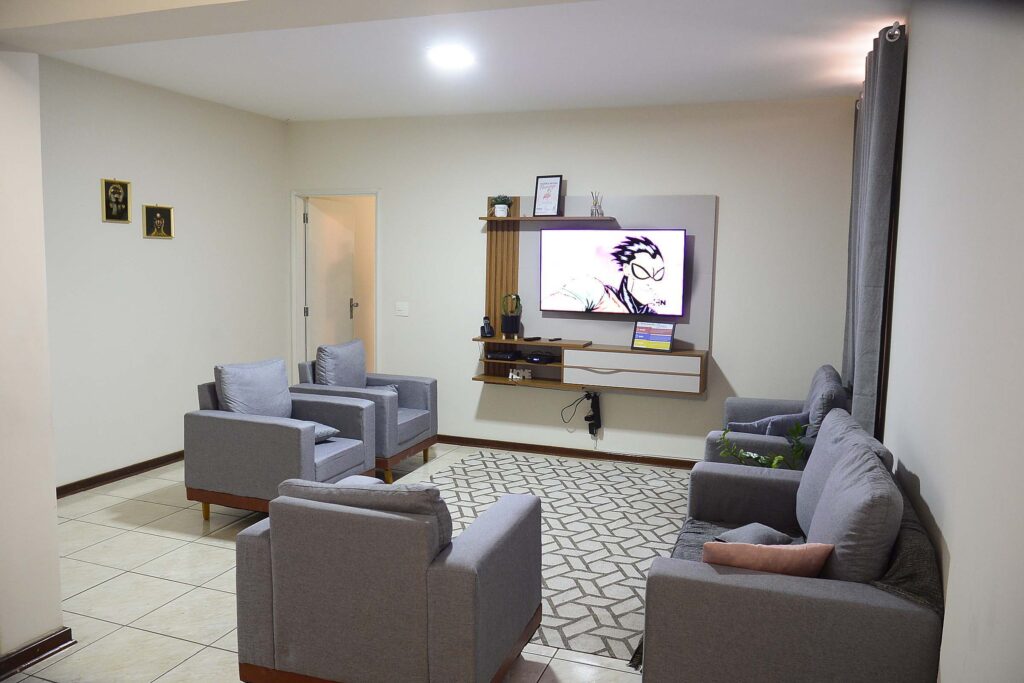 A imagem mostra uma sala de convivência da Residência Inclusiva, ampla, organizada e bem iluminada. O espaço conta com sofás e poltronas, tapete, plantas decorativas e uma televisão instalada em um painel de madeira. Ao fundo, há quadros decorativos e uma porta de acesso a outro ambiente.