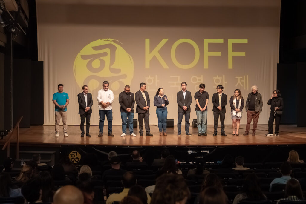 A imagem mostra um grupo de doze pessoas em pé sobre um palco, diante de uma plateia. No fundo, há uma grande projeção com o logotipo circular amarelo e o texto “KOFF” acompanhado de caracteres coreanos e da frase “Korean Film Festival”.
