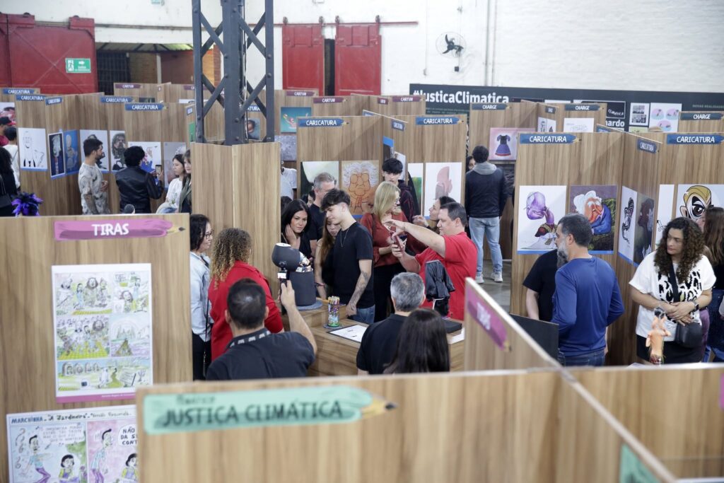 Esta imagem mostra uma exposição de artes gráficas, parecida com uma feira de quadrinhos ou cartuns, realizada em um amplo espaço com teto alto e vigas aparentes. Aqui estão os detalhes principais: O Ambiente: O espaço está organizado com divisórias de madeira que criam "estandes" individuais, onde várias obras estão expostas. Há sinalizações no topo das divisórias indicando as categorias das obras, como "Caricatura", "Tiras" e "Charge". O Público: Há várias pessoas circulando pelo local, observando os quadros e conversando. No centro, nota-se um grupo mais concentrado ao redor de uma mesa onde parece estar acontecendo algum tipo de demonstração artística ou venda; um homem de camiseta vermelha está ali segurando um celular, possivelmente registrando algo. Temática: Além das artes expostas, há uma sinalização destacada ao fundo e na parte frontal inferior com os dizeres "Justiça Climática", sugerindo que a exposição aborda temas sociais e ambientais através do humor gráfico. Detalhes Visuais: À esquerda, um cartaz expõe "Tiras" com personagens. Do lado direito, há várias caricaturas coloridas de personalidades famosas ou figuras estilizadas. O clima é de um evento cultural bem frequentado, onde as pessoas interagem com as artes e entre si.