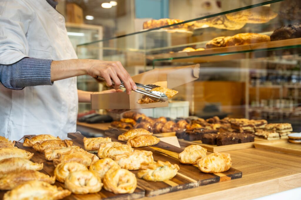 A imagem mostra uma padaria ou confeitaria com um balcão repleto de produtos assados. Uma pessoa usando avental branco utiliza uma pinça para colocar um salgado dourado dentro de uma caixa de papelão. Sobre as tábuas de madeira, há muitos folhados, empadas, croissants e outros pães e doces. Ao fundo, uma vitrine de vidro exibe mais produtos de panificação, iluminados por uma luz quente e acolhedora.