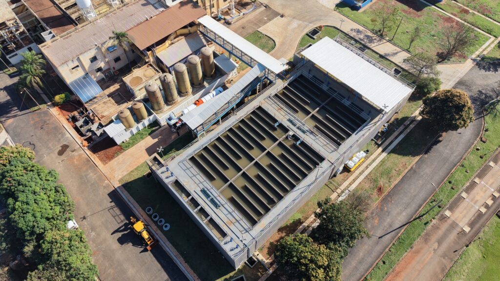 Na imagem, observa-se uma vista aérea de uma instalação industrial dedicada ao tratamento de água. No centro, destacam-se grandes tanques retangulares com divisórias paralelas, provavelmente usados em processos de sedimentação ou filtração. Ao redor, há reservatórios cilíndricos, tubulações e pequenos edifícios que abrigam equipamentos e sistemas de controle. O espaço é bem organizado, delimitado por ruas pavimentadas, áreas verdes e árvores, transmitindo a ideia de uma estrutura planejada para garantir eficiência e sustentabilidade na gestão dos recursos hídricos.