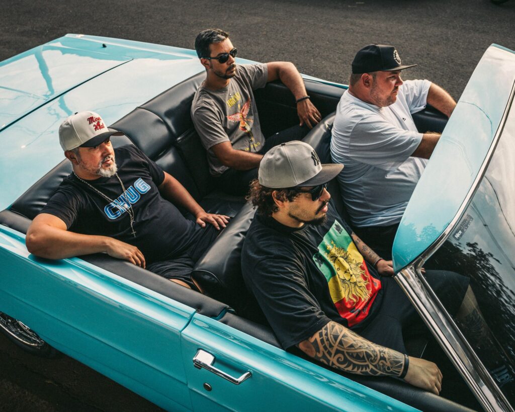A imagem mostra quatro homens dentro de um carro conversível azul-petróleo, visto de cima e ligeiramente de lado. O carro tem bancos pretos e o topo está abaixado. O asfalto escuro cobre o fundo. À esquerda, na frente, está um homem com barba grisalha usando um boné branco e cinza com a inscrição "Mono Heat" em vermelho. Ele veste uma camiseta preta com a palavra "CHUG" em letras azuis e um colar com um pingente em forma de cruz. Ele está encostado no banco, com o braço esquerdo repousando na porta. Atrás dele, no banco do motorista, está um homem com um boné preto e uma camiseta branca. Ele está olhando para a frente e para a direita. No banco do passageiro da frente, à direita, está um homem com óculos escuros e um boné cinza com o logo do New York Yankees. Ele veste uma camiseta preta com uma estampa vibrante de um leão com crina amarela, emoldurado por faixas verdes, amarelas e vermelhas, remetendo às cores do movimento rastafári. Seus braços exibem tatuagens detalhadas em preto. Ele parece estar olhando para a frente. Atrás dele, no banco do passageiro de trás, está um homem com óculos escuros e uma camiseta cinza. A camiseta tem uma estampa com a figura de um anjo alado e a palavra "NIRVANA". Ele está olhando para a esquerda e para a frente, com uma pulseira escura no pulso. A luz do sol reflete na pintura brilhante do carro e em seu para-brisa. A composição é dinâmica, capturando um momento casual e descontraído entre os homens no carro. As cores predominantes são o azul vibrante do carro, o preto dos bancos e das roupas, e os tons neutros dos bonés e de algumas camisetas, contrastando com as estampas coloridas.