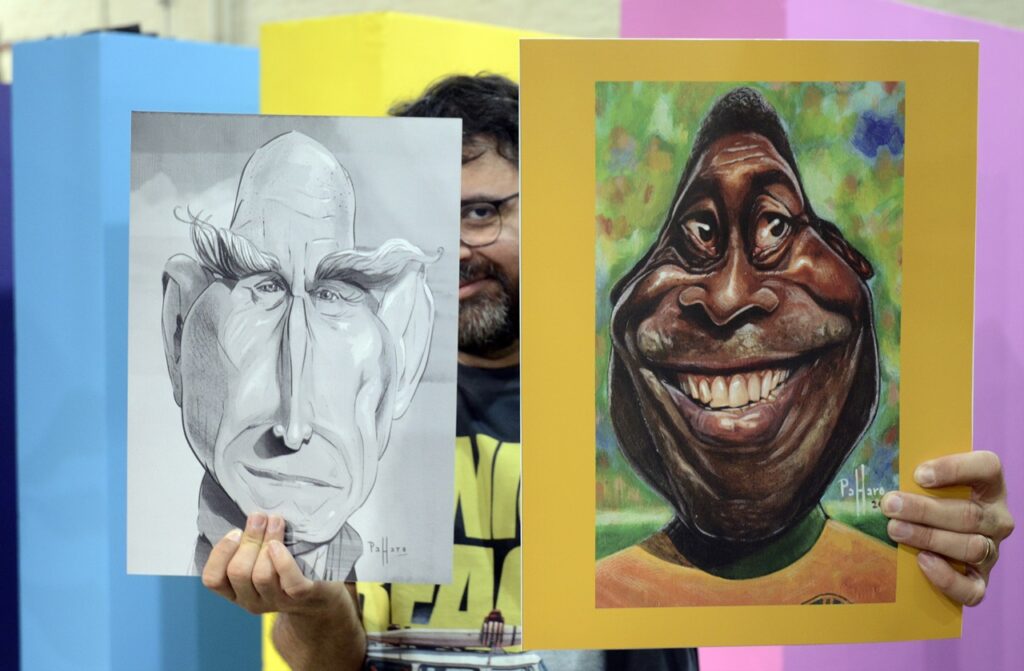 A imagem mostra um homem segurando duas caricaturas lado a lado, cobrindo parcialmente o rosto. À esquerda, há um desenho em preto e branco de um homem com traços exagerados — rosto alongado, orelhas grandes e sobrancelhas espessas e curvas. À direita, uma caricatura colorida de um homem sorridente, com dentes grandes e expressão alegre, sobre um fundo verde e colorido.