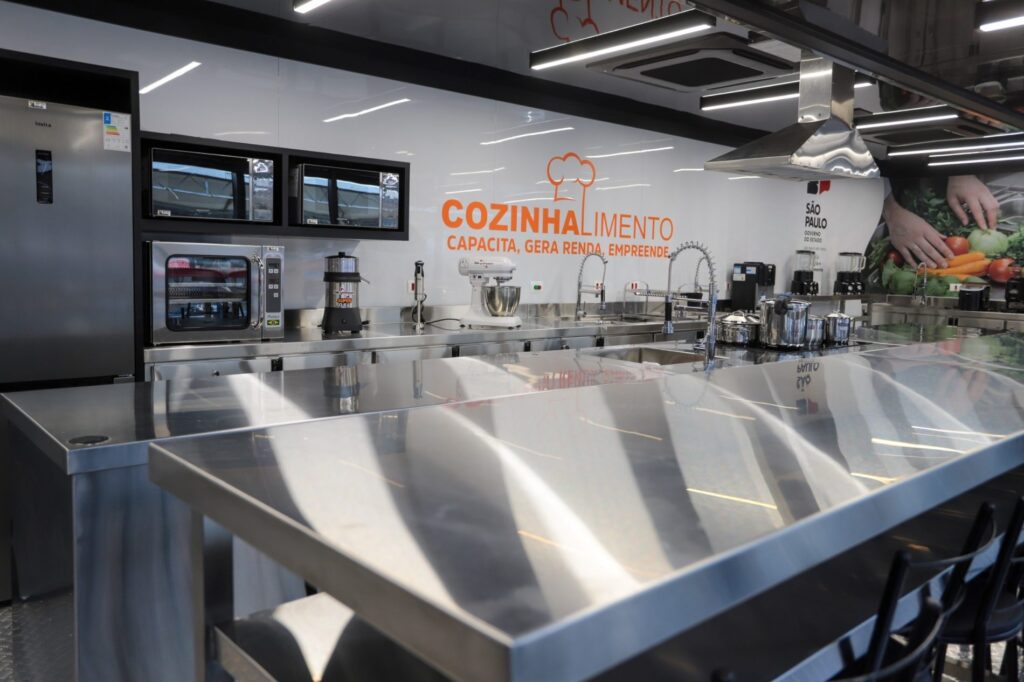 A imagem mostra o interior de uma cozinha profissional, moderna e muito bem equipada, provavelmente instalada em uma unidade móvel de capacitação. No centro da parede aparece o texto “Cozinhalimento — Capacita, gera renda, empreende”, junto ao logotipo do Governo do Estado de São Paulo. Há bancadas amplas de aço inoxidável, pias com torneiras altas, fornos, micro-ondas, liquidificadores, batedeira industrial, panelas e outros utensílios. No teto, vê-se um grande sistema de exaustão e várias luminárias. As superfícies metálicas refletem a iluminação, deixando o ambiente claro, organizado e adequado para aulas práticas de culinária.