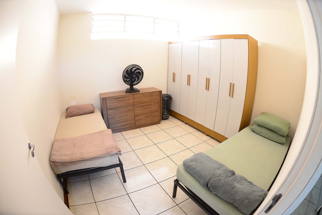 A imagem mostra um quarto simples e bem organizado, com duas camas de solteiro posicionadas em lados opostos. A cama à esquerda tem roupa de cama bege e um travesseiro marrom, enquanto a da direita tem lençóis verdes e uma manta cinza dobrada aos pés. No centro da parede do fundo há uma cômoda de madeira com quatro gavetas, sobre a qual está colocado um ventilador preto. Ao lado, há um cesto de roupa e, à direita, um guarda-roupa grande com portas brancas e puxadores de madeira.