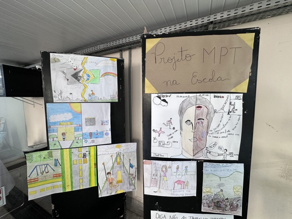A imagem mostra uma exposição escolar montada em painéis pretos, com o título “Projeto MPT na Escola”. Os trabalhos abordam o tema do trabalho infantil, representando situações como crianças trabalhando, a importância da escola, brincadeiras, desigualdade social e os direitos da infância. Há desenhos coloridos e em preto e branco, incluindo um rosto dividido entre uma infância protegida e outra marcada pelo trabalho. O ambiente parece ser o corredor ou uma área interna de uma escola.