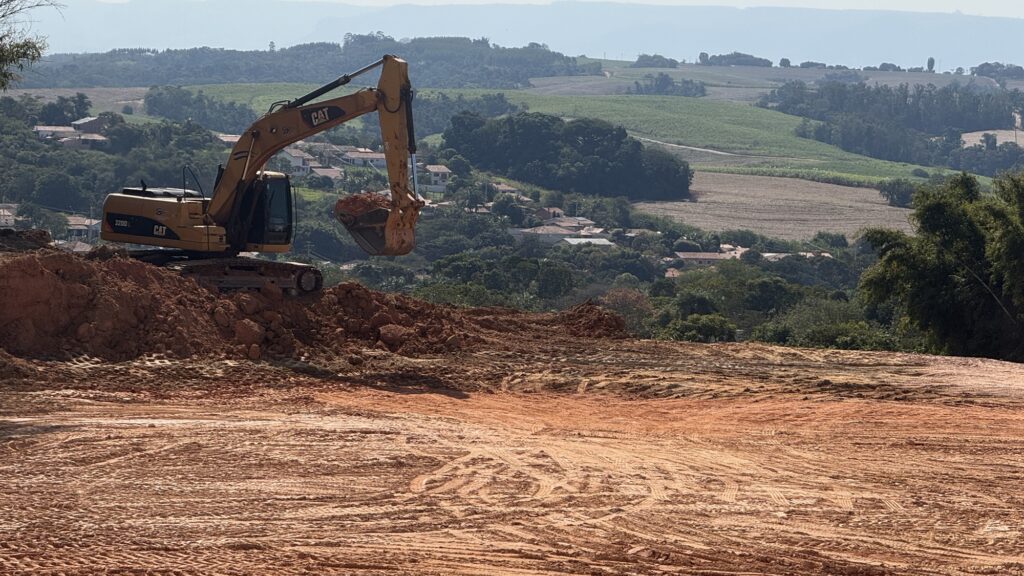A imagem mostra um canteiro de obras em uma encosta, onde uma escavadeira amarela modelo CAT 320D está em plena atividade, levantando terra com sua pá. O solo, de tom avermelhado, contrasta com o cenário ao fundo: campos verdes, casas espalhadas e colinas cobertas por árvores que se estendem até o horizonte. No primeiro plano, o terreno recém-nivelado exibe marcas de pneus e trilhas deixadas pelas máquinas pesadas, reforçando a ideia de transformação do espaço.