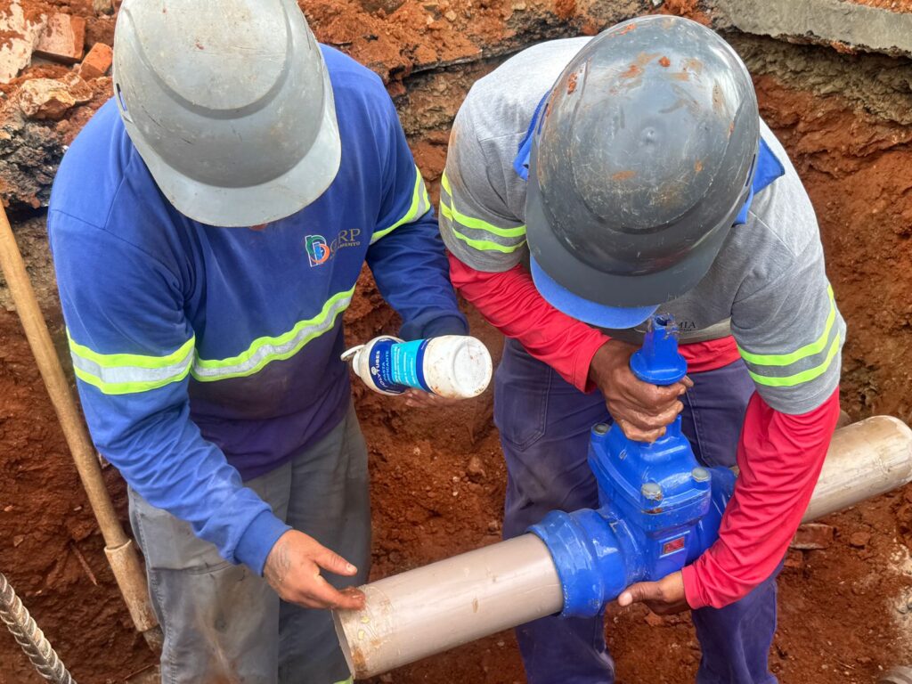 A imagem mostra dois trabalhadores da construção civil em um fosso aberto com terra avermelhada ao redor. Ambos usam capacetes de segurança, um cinza claro e o outro cinza escuro. O trabalhador da esquerda veste uma camisa azul de manga longa com detalhes refletivos em verde limão nas mangas e no peito, além de um logotipo em branco e laranja. Ele está segurando um tubo bege com a mão direita, enquanto com a mão esquerda segura um tubo de lubrificante branco com rótulo azul e branco escrito "OXITUBES LUBRIFICANTE". O trabalhador da direita veste uma camisa cinza de manga longa com detalhes refletivos em verde limão nas mangas, e por baixo, uma manga vermelha visível. Ele usa calças azuis escuras. Ele está concentrado em uma válvula azul brilhante conectada a um tubo bege horizontal, segurando-a com as duas mãos. O ambiente é de uma obra, com a terra exposta e alguns tijolos quebrados ao fundo. A iluminação parece ser natural, vindo de cima, criando sombras suaves. A composição foca na ação dos trabalhadores e nos materiais que estão manuseando, transmitindo a ideia de trabalho em equipe e execução de uma tarefa específica em uma obra de infraestrutura. As cores predominantes são os tons terrosos da terra, o azul das roupas e da válvula, e os detalhes refletivos que se destacam.