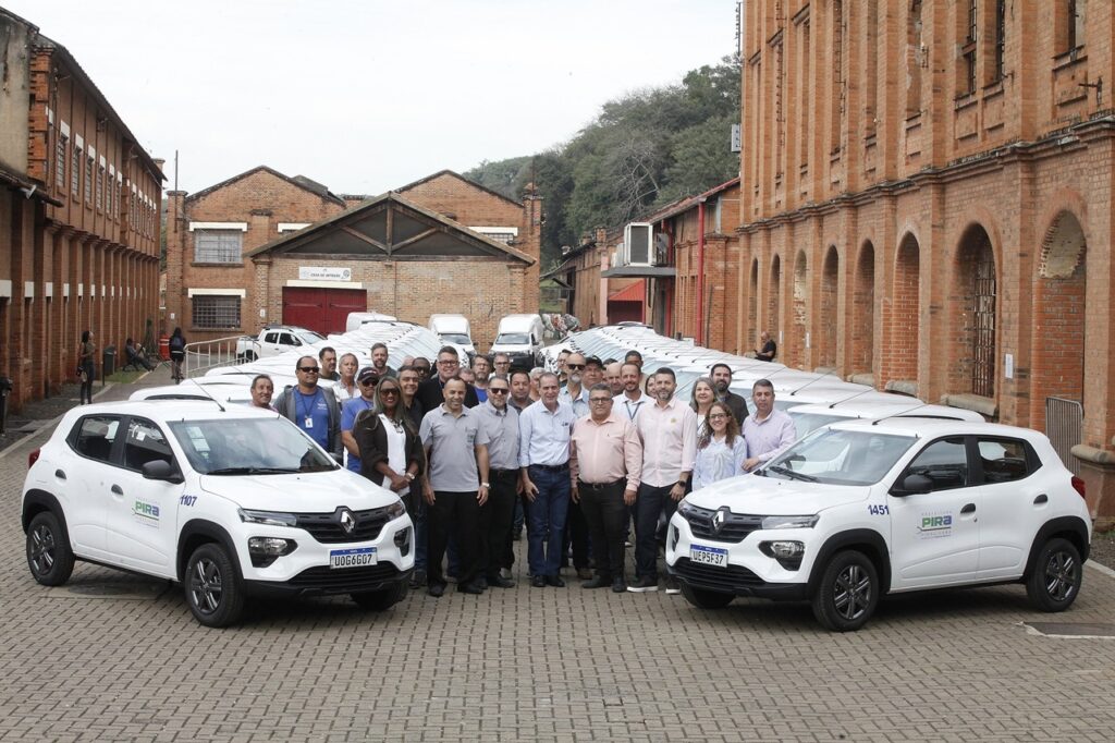 Esta foto registra um momento oficial de entrega de uma nova frota de veículos para a Prefeitura de Piracicaba. O grupo: Uma grande equipe posa para a foto em frente à frota. No centro, destacam-se algumas autoridades e funcionários, muitos sorrindo, posando em um formato de "V" aberto. Entre eles, há uma diversidade de trajes, desde camisas polo e sociais até roupas casuais. A frota: Vários carros brancos, modelo Renault Kwid, estão estacionados em duas fileiras longas que se estendem para o fundo, criando uma linha de perspectiva. Nas laterais dos carros, pode-se ler "Prefeitura Pira" e a numeração da frota (ex: 1107, 1451). O cenário: O local é um pátio pavimentado com blocos de concreto, situado entre grandes edifícios de tijolos aparentes, caracterizados por arcos nas janelas e uma arquitetura industrial antiga. Ao fundo, ao centro, é possível ver uma placa com os dizeres "Casa do Artesão", o que situa o local dentro de um complexo histórico ou de cultura. A atmosfera é de um evento institucional de grande porte, com clima de conquista e renovação.