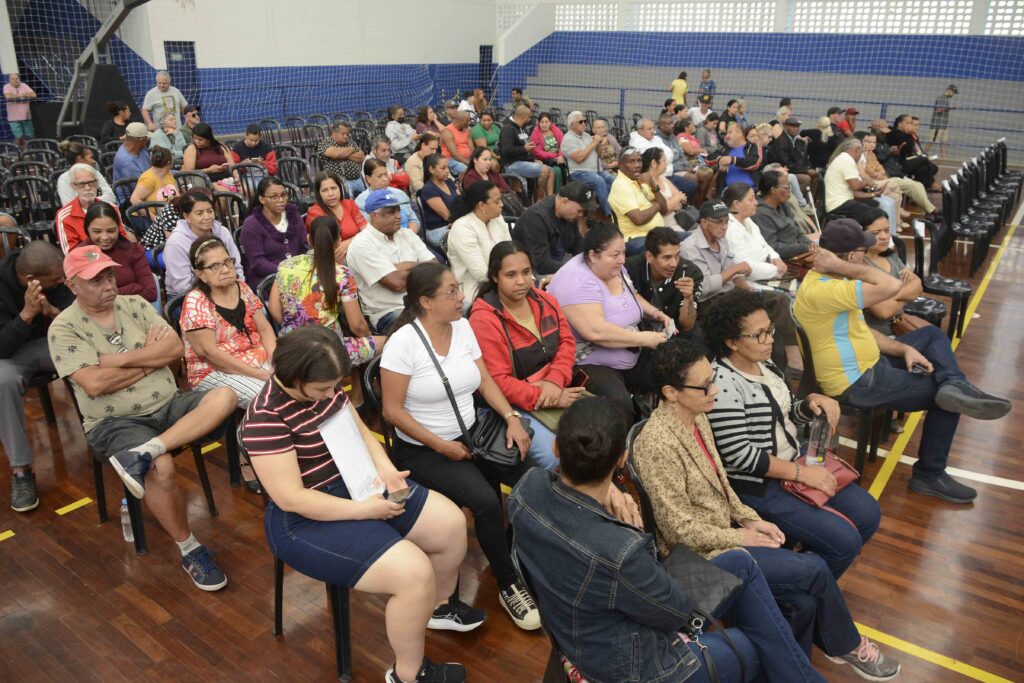 A imagem mostra um grupo grande de pessoas sentadas em cadeiras dentro de um ginásio coberto, aguardando ou participando de um evento comunitário. O piso é de madeira e as paredes têm detalhes em azul, com uma rede de proteção típica de quadras esportivas. O público é diverso, composto por homens e mulheres de diferentes idades, muitos atentos e conversando entre si. O ambiente transmite um clima de reunião pública ou cerimônia social.