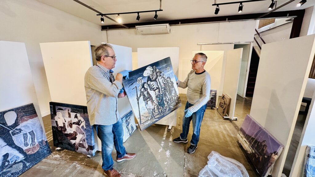 A cena mostra dois homens montando uma exposição de arte em um espaço de galeria. Ação: Eles seguram uma pintura que retrata um grupo de pessoas — parece uma obra que remete ao estilo de Cândido Portinari — e a posicionam cuidadosamente contra um painel divisório branco. Ambos usam luvas azuis para não danificar a tela. Ambiente: É um local bem organizado, com várias outras pinturas espalhadas pelo chão, algumas ainda protegidas por plástico bolha. O piso é de cimento queimado e o teto possui iluminação de trilho. Clima: Transmite um momento de bastidores, com foco, precisão e muito cuidado com as obras de arte.