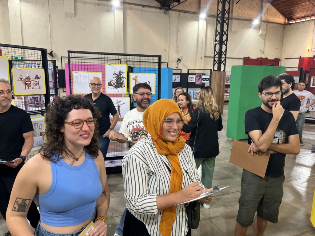 A imagem mostra um grupo de pessoas reunido em uma exposição de arte, dentro de um amplo galpão com teto alto e estrutura metálica. Em primeiro plano, há duas mulheres: uma usa blusa azul e óculos, e a outra veste um lenço mostarda, camisa listrada e também usa óculos. Ao redor delas, várias pessoas observam as obras e conversam; algumas seguram pranchetas. Ao fundo, painéis coloridos exibem ilustrações, desenhos e fotografias, incluindo personagens fantásticos e animais. O ambiente é bem iluminado e tem piso de concreto, com uma grande estrutura verde à direita.