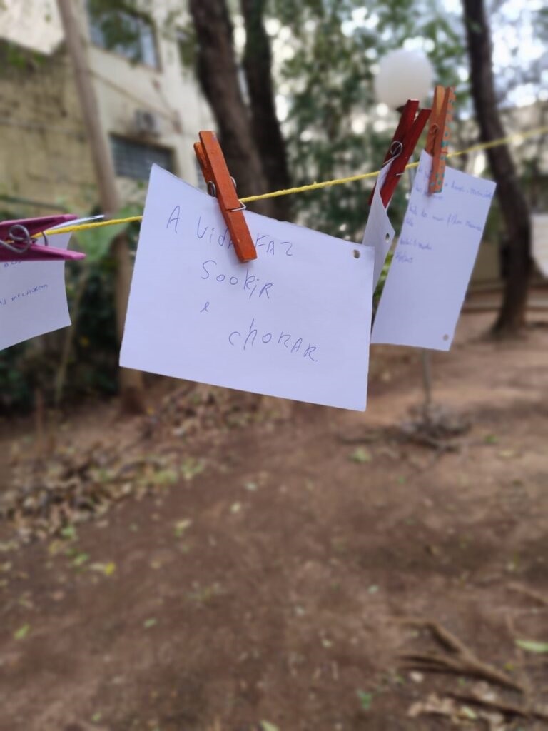 A imagem mostra vários cartões de papel pendurados em um varal com pregadores coloridos, em uma área externa arborizada. O cartão central traz a frase manuscrita: “A vida faz sorrir e chorar”. Ao fundo, aparecem árvores, o chão de terra e parte de uma construção, todos desfocados.