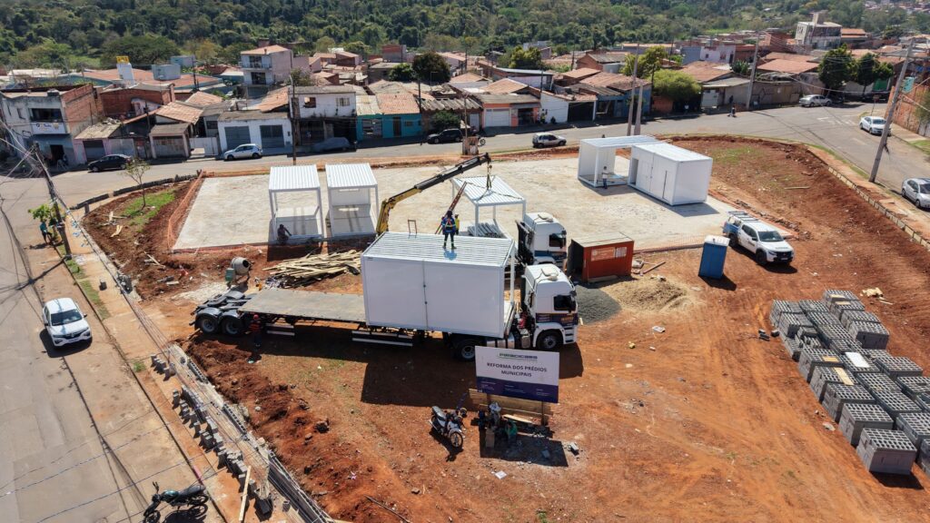 Esta fotografia aérea captura uma cena de construção urbana onde trabalhadores utilizam um caminhão equipado com guindaste para posicionar estruturas modulares brancas em um terreno de terra batida. A composição da cena é vista de uma perspectiva aérea, apresentando um canteiro de obras em um terreno de esquina delimitado por ruas pavimentadas e casas residenciais ao fundo. A ação central gira em torno do posicionamento de módulos pré-fabricados, com um caminhão centralizado carregando um módulo, enquanto outros já estão instalados sobre uma base de concreto. A disposição dos elementos cria um fluxo de trabalho logístico, com pilhas de blocos de concreto organizadas à direita e o maquinário operando ativamente. A profundidade é dada pela transição entre o canteiro de obras em primeiro plano e a infraestrutura urbana ao fundo. O assunto principal é a atividade de construção. Vemos pelo menos um trabalhador posicionado no topo do módulo branco sobre o caminhão, coordenando a operação. O caminhão de carga, de cor branca com cabine robusta, é o ponto focal da movimentação. Pessoas vestindo uniformes de trabalho são visíveis perto das estruturas, garantindo que o encaixe dos módulos seja preciso. A presença de um guindaste braço mecânico estendido sugere um momento de precisão técnica na montagem das estruturas pré-moldadas, que possuem paredes lisas e brancas, assemelhando-se a contêineres funcionais. A imagem é uma fotografia digital capturada provavelmente por um drone, apresentando uma qualidade técnica nítida com foco profundo que mantém tanto o primeiro plano quanto as casas ao fundo visíveis. O estilo é documental e informativo, comum em registros de engenharia ou acompanhamento de obras públicas. A iluminação é natural e direta, vinda de um dia ensolarado, o que resulta em sombras curtas sob os módulos e veículos, enfatizando o volume e a tridimensionalidade das formas geométricas simples que compõem o canteiro. O cenário é um ambiente urbano em fase de desenvolvimento ou reforma, com um terreno marcado por terra avermelhada típica de regiões tropicais, situado em uma área residencial com casas de alvenaria próximas. A luz solar forte e direta cria uma atmosfera de trabalho diurno e produtivo. Os elementos ambientais, como a placa de sinalização da obra em destaque no centro e as pilhas organizadas de materiais de construção, reforçam a sensação de um projeto em andamento. O contraste entre a terra vermelha exposta e as estruturas brancas brilhantes confere à cena uma estética industrial e organizada, típica de projetos de infraestrutura municipal.