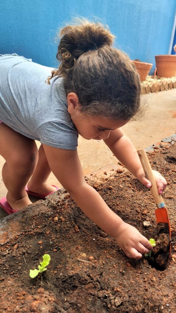 A imagem mostra uma criança agachada, plantando mudas em um canteiro de terra. Ela usa uma camiseta cinza, shorts e chinelos cor-de-rosa, além de prender o cabelo em um coque. Com uma pequena pá de jardinagem, a criança mexe na terra ao redor de uma muda recém-plantada. Outra muda aparece mais à frente no canteiro. Ao fundo, há uma parede azul, vasos de plantas e um espaço externo ensolarado.