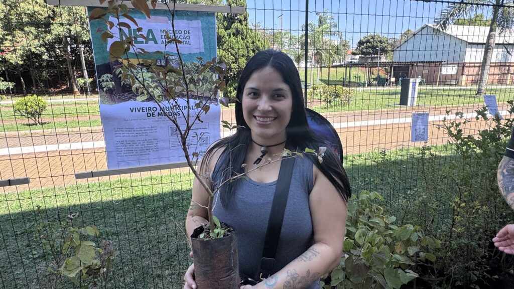 Na foto, vemos uma pessoa em um espaço ao ar livre, como um parque ou jardim, segurando uma muda de árvore em um saco plástico de plantio. Atrás dela há uma cerca de arame com um cartaz que traz os dizeres “NEA Núcleo de Educação Ambiental” e “Viveiro Municipal de Mudas”, indicando que a cena está ligada a uma iniciativa de educação ambiental e reflorestamento comunitário. O entorno mostra grama verde, árvores, um caminho pavimentado e um prédio de telhado branco ao fundo.