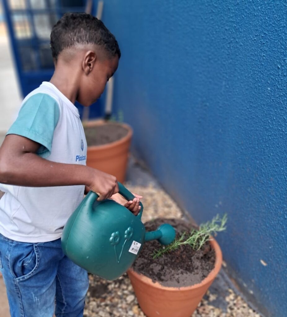 A imagem mostra um menino regando uma planta em um vaso de barro. Ele segura um regador verde com as duas mãos e direciona a água para a terra. O menino veste uma camiseta branca com mangas azul-turquesa, com o logotipo da Prefeitura de Piracicaba, e uma bermuda jeans. Ao fundo, há uma parede azul, outros vasos e um corredor externo com pedrinhas no chão.