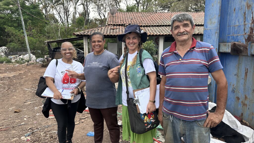 A imagem mostra quatro adultos sorrindo e posando para uma foto em uma área externa. Duas mulheres seguram pranchetas com formulários; uma delas usa camiseta com a mensagem “Desconfiar da tuberculose”, enquanto outra veste um colete com identificação da Prefeitura de Piracicaba. Ao lado, há uma mulher usando chapéu e mochila, e um homem com camisa polo listrada. Eles estão diante de uma casa simples com telhado de telhas, vegetação ao redor e um portão metálico azul enferrujado. O chão tem terra, materiais espalhados e sacos, sugerindo uma visita de campo ou ação de saúde comunitária.