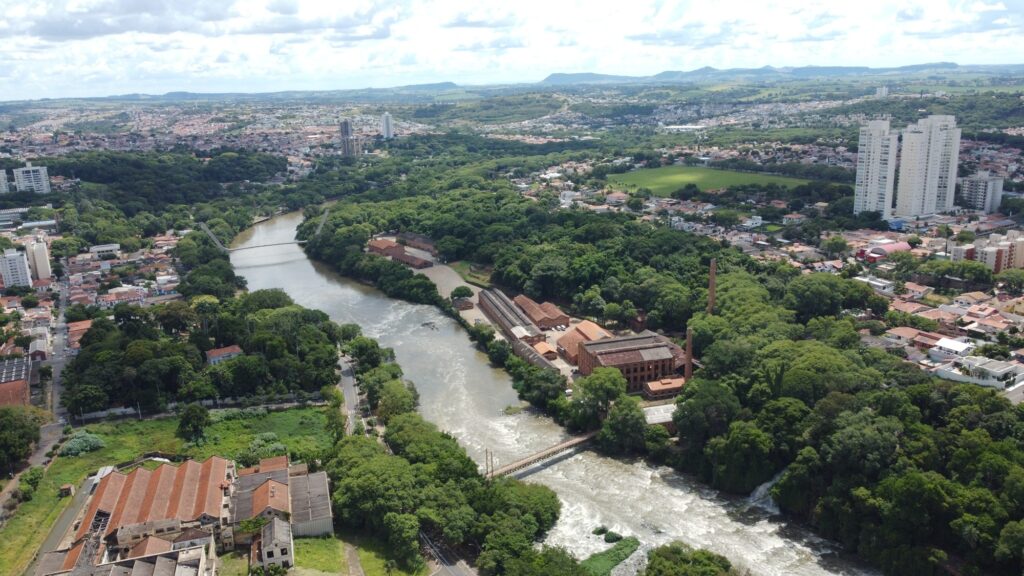 A imagem mostra uma vista aérea de uma cidade cortada por um rio que serpenteia entre áreas densamente arborizadas. Nas margens, destacam-se grandes construções de tijolos com telhados vermelhos, lembrando antigas fábricas ou moinhos, além de uma chaminé alta que reforça o caráter industrial do local. Um passarela atravessa o rio, conectando os dois lados. Ao redor, há bairros residenciais com casas e prédios, e mais ao fundo surgem edifícios modernos que se estendem até colinas distantes.