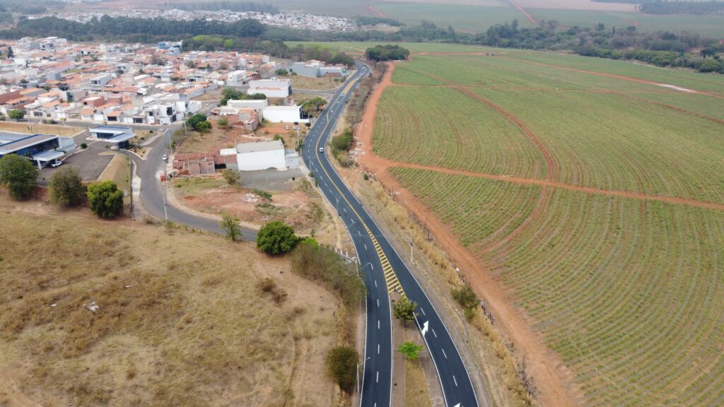 A imagem aérea mostra uma área urbana na transição para a zona rural. À esquerda, há um bairro residencial com casas de telhados alaranjados, ruas pavimentadas e alguns edifícios comerciais. Uma estrada recém-pavimentada, com faixas amarelas e brancas, curvas e sinalização, atravessa a cena e separa o bairro das extensas plantações à direita. Veem-se alguns veículos circulando pela via. Ao fundo, há mais casas, áreas arborizadas e grandes campos agrícolas com fileiras regulares de cultivo, sob um céu nublado.