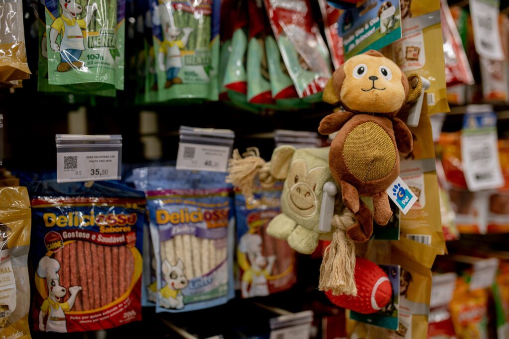 Uma prateleira de um pet shop exibe uma variedade de petiscos embalados e brinquedos de pelúcia, criando um ambiente de compras organizado que apela diretamente aos donos de animais de estimação. A cena é composta por uma estante de varejo onde diversos pacotes coloridos de guloseimas caninas estão pendurados lado a lado, ocupando a maior parte do fundo. Em destaque na parte frontal direita, há um agrupamento de brinquedos de pelúcia que servem como ponto focal, criando um contraste interessante entre a rigidez dos pacotes plásticos e a textura macia dos bichinhos. A composição segue um padrão típico de exposição comercial, com etiquetas de preços e códigos QR visíveis, organizando o espaço visual e orientando o olhar do observador através das diferentes seções de produtos para animais. O sujeito principal no centro da atenção é um brinquedo de pelúcia em formato de macaco, com tons de marrom e bege, apresentando uma expressão simpática com olhos grandes e um sorriso bordado. Logo abaixo dele, nota-se uma parte de um brinquedo de pelúcia esverdeado com feições geométricas e uma bola de borracha vermelha texturizada, que adiciona um toque vibrante à composição. As embalagens de petiscos ao fundo são predominantemente azul e vermelha, trazendo ilustrações de cães animados como mascotes, com textos e marcas bem definidos, mantendo uma estética comercial vibrante e informativa, característica comum em corredores de lojas de produtos para pets. A fotografia utiliza uma técnica de foco seletivo, mantendo os brinquedos de pelúcia em primeiro plano com clareza nítida, enquanto os produtos dispostos nas prateleiras mais distantes apresentam um efeito de desfoque suave, conhecido como bokeh. Esta escolha técnica enfatiza a tridimensionalidade dos objetos em destaque e direciona o foco do espectador para a variedade de itens disponíveis para entretenimento dos animais. A luz é artificial, vinda de cima, típica de interiores comerciais, o que gera sombras sutis e realça as texturas do tecido dos bichos de pelúcia e o brilho do plástico das embalagens, conferindo um ar limpo e acolhedor ao ambiente da loja. O cenário é o corredor de uma loja de suprimentos para animais, ambientado de forma funcional para maximizar a visibilidade dos produtos. A atmosfera é iluminada e organizada, transmitindo uma sensação de abundância e cuidado voltado para o bem-estar animal. As cores predominantes são os tons terrosos dos brinquedos contrastando com as embalagens vibrantes, o que ajuda a criar uma experiência visual organizada para o consumidor. O ambiente reflete um espaço de conveniência moderno, onde a disposição estratégica dos itens, desde os alimentos até os acessórios de lazer, convida à interação e à exploração do cliente, mantendo um clima de otimismo e utilidade prática.