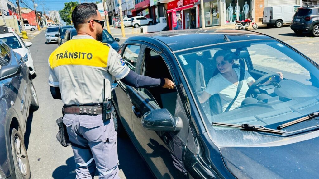 A imagem mostra um agente de trânsito abordando uma motorista em uma rua movimentada. O agente veste uniforme com a palavra “TRÂNSITO” nas costas, usa óculos escuros e carrega um rádio comunicador e outros equipamentos no cinto. A motorista está sentada em um carro escuro, com o cinto de segurança afivelado, enquanto conversa com o agente pela janela aberta. Ao redor, há vários veículos estacionados e em circulação, lojas, motocicletas e prédios comerciais. O dia está ensolarado.