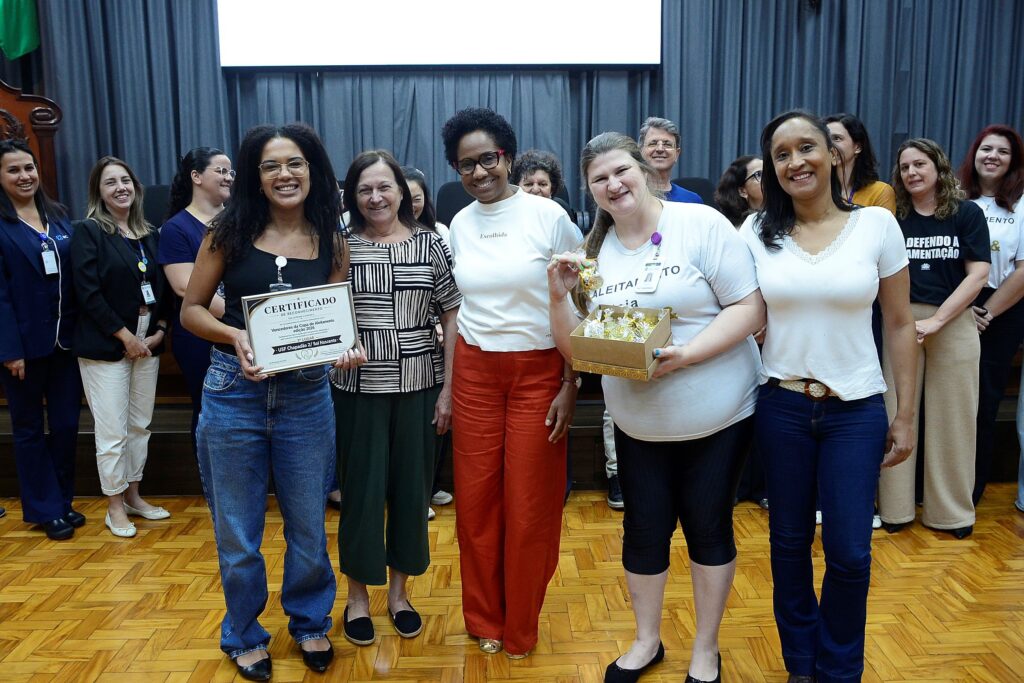A imagem mostra um grupo de pessoas, na maioria mulheres, celebrando uma conquista em um ambiente que parece ser um auditório ou sala de conferências. O clima é de felicidade e reconhecimento profissional. Aqui estão os detalhes principais: A Celebração: O grupo está reunido para comemorar uma premiação. No centro, uma das participantes segura um "Certificado de Reconhecimento", que identifica os ganhadores do 3º lugar na "Copa do Aleitamento, edição 2026", especificamente para a "USF Chapadão 2/ Sol Nascente". Os Detalhes: Ao lado da pessoa com o certificado, outra participante segura uma caixa contendo pequenos mimos (parecem doces embrulhados em papel dourado). O Ambiente: Elas estão posicionadas sobre um piso de madeira (parquet), com cortinas cinzas ao fundo, em um cenário típico de eventos institucionais ou de saúde pública. O Grupo: As pessoas estão sorridentes, posando para uma foto de equipe, vestindo roupas casuais e profissionais, como camisetas, jeans e blazers.