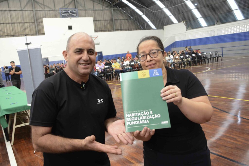A imagem mostra duas pessoas em um ginásio coberto, sorrindo e segurando um documento verde da Secretaria Municipal de Habitação e Regularização Fundiária. Ao fundo, há várias pessoas sentadas, indicando que o local abriga um evento público ou cerimônia de entrega de documentos. O ambiente é organizado, com mesas cobertas por toalhas verdes e uma atmosfera de participação comunitária.