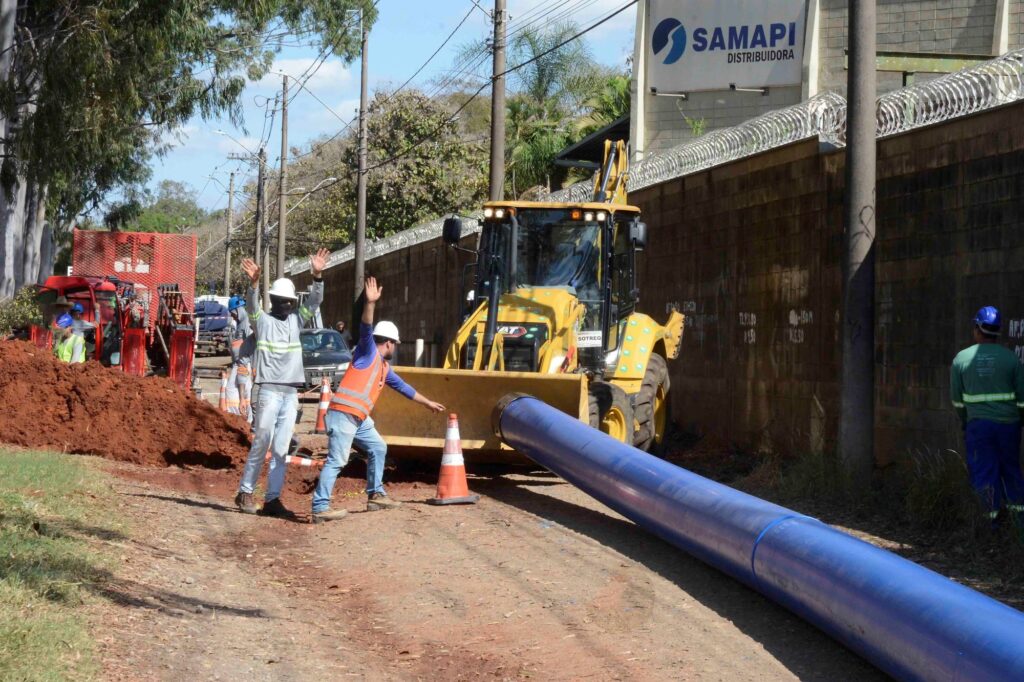 A imagem mostra uma obra de manutenção em uma área externa, ao lado de um muro com arame farpado. Vários trabalhadores usam capacetes, coletes refletivos e equipamentos de segurança, enquanto orientam a operação. No centro, há uma retroescavadeira amarela e, à esquerda, máquinas e veículos de apoio. Um grande tubo azul está sendo instalado ou reparado no solo, próximo a uma vala aberta e a cones de sinalização. No alto, aparece a placa “SAMAPI Distribuidora”. Ao fundo, há árvores, postes e muitos fios elétricos.