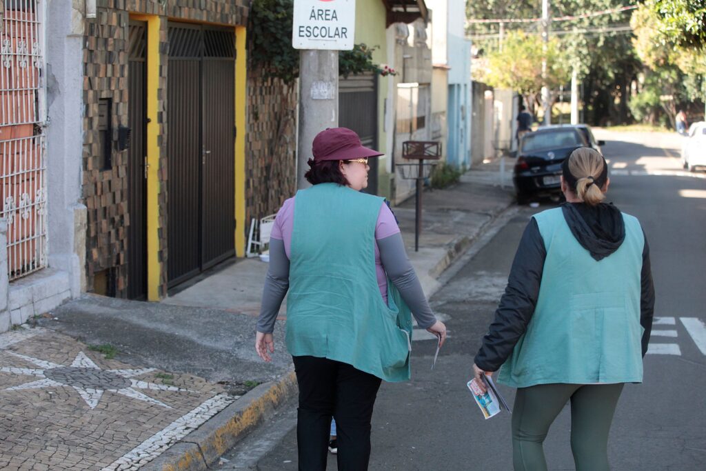 Duas mulheres uniformizadas com coletes verdes caminham por uma rua residencial tranquila, sob a luz do dia, em uma cena que sugere trabalho de campo ou uma atividade comunitária externa. A composição da cena é focada nas costas das duas mulheres, que caminham lado a lado em direção ao fundo da rua, estabelecendo um movimento constante de afastamento em relação ao observador. Elas estão posicionadas no centro da via, com elementos arquitetônicos de casas à esquerda e a perspectiva da rua estendendo-se à direita, criando uma sensação de profundidade e direcionamento. A interação é silenciosa, com ambas concentradas na caminhada e segurando papéis, reforçando a ideia de uma tarefa em execução. As duas sujeitas principais são mulheres adultas vestidas de maneira funcional para atividades externas. A mulher à esquerda veste uma blusa de mangas compridas em tom rosado, calças pretas, um boné bordô e um colete verde característico. A mulher à direita usa uma jaqueta preta com capuz sob o mesmo modelo de colete verde, calças estilo legging em tom esverdeado e está com o cabelo preso em um rabo de cavalo. Ambas carregam pequenos impressos ou folhetos nas mãos, sugerindo uma função de distribuição ou coleta de informações. Esta é uma fotografia digital capturada com foco nítido nos sujeitos em primeiro plano, utilizando uma lente que mantém um equilíbrio realista entre os elementos urbanos e a figura humana. A técnica de captura é documental, com cores naturais e uma iluminação difusa que elimina sombras fortes, conferindo um aspecto sóbrio e cotidiano à imagem. A escolha de enquadramento captura a cena de forma aberta, permitindo a visibilidade da sinalização urbana e das texturas das calçadas e fachadas das casas próximas. O cenário é uma rua urbana de perfil residencial, com casas de fachadas simples e muros de alvenaria. Uma placa de trânsito indicando "ÁREA ESCOLAR" está fixada em um poste central, fornecendo um contexto geográfico e situacional sobre a vizinhança. A iluminação é natural e suave, típica de um dia nublado ou de um período do dia onde o sol não incide diretamente, o que proporciona uma atmosfera calma e serena. O ambiente circundante é composto por elementos típicos de uma zona suburbana, com árvores ao fundo e um carro estacionado, criando um clima de normalidade e rotina.
