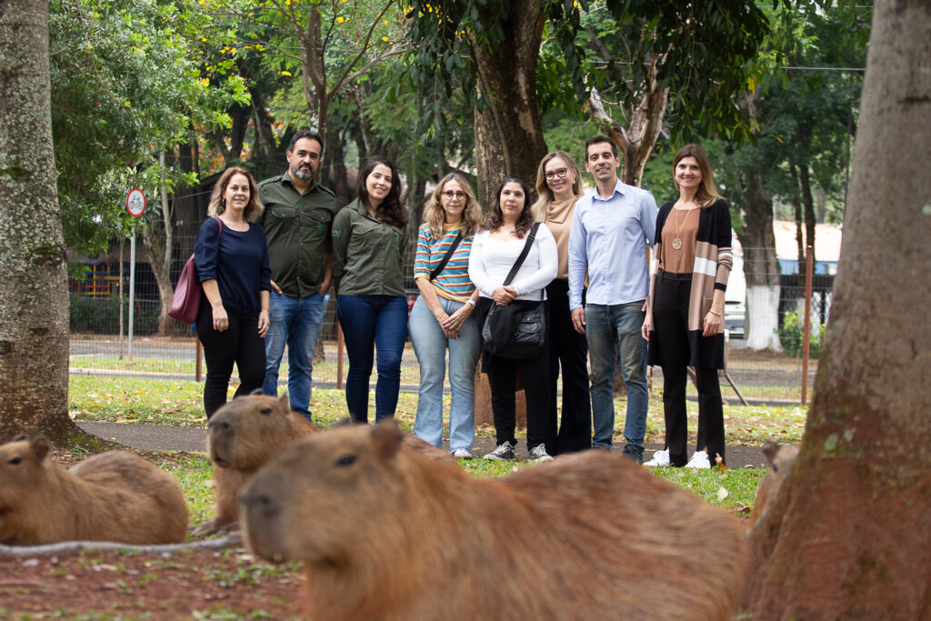 Uma fotografia ao ar livre captura um grupo diversificado de oito adultos posando casualmente em um parque arborizado, enquanto várias capivaras repousam tranquilamente no primeiro plano, criando um contraste fascinante. A composição é organizada em duas camadas distintas, com as capivaras ocupando o primeiro plano inferior, criando um elemento de interesse natural, enquanto o grupo de pessoas está alinhado horizontalmente na área gramada do meio. O enquadramento é amplo, aproveitando as árvores laterais para emoldurar a cena. A interação é de uma observação pacífica e estática; as pessoas olham diretamente para a câmera, enquanto os animais permanecem relaxados, ignorando a presença humana, o que sugere um ambiente público onde ambos os grupos coexistem habitualmente. O grupo é composto por seis mulheres e dois homens, todos vestidos com roupas casuais do dia a dia, como jeans, camisas de botão, blusas e cardigãs, apresentando uma paleta de cores sóbrias e terrosas que harmonizam com o ambiente. Suas expressões são serenas e sorridentes, indicando uma ocasião de lazer ou uma reunião informal. As capivaras, em destaque no primeiro plano, possuem pelagem densa e áspera em tons de marrom, com características físicas robustas, focinhos quadrados e olhos pequenos e alertas, típicos de seu estado de descanso vigilante. Esta imagem é uma fotografia digital de estilo documental, caracterizada pela sua honestidade visual e foco nítido nos sujeitos humanos, com uma profundidade de campo que permite que tanto o grupo quanto as capivaras sejam claramente identificáveis. A técnica privilegia a luz natural e uma abordagem sem retoques excessivos, mantendo as texturas naturais da grama, da casca das árvores e dos pelos dos animais. O equilíbrio entre o foco no grupo e a presença deliberada dos animais em primeiro plano sugere uma escolha criativa que busca documentar a convivência urbana com a fauna silvestre. O cenário é um parque público bem arborizado, com troncos de árvores grandes servindo como moldura lateral, e uma iluminação natural difusa, típica de um dia nublado ou de um ambiente sob sombra densa de copas. A luz suave evita sombras duras no rosto das pessoas e destaca os detalhes da pelagem das capivaras. O ambiente, que inclui uma estrada pavimentada ao fundo e uma grade de proteção, reforça a atmosfera de um espaço de lazer urbano. O clima geral é de tranquilidade e integração entre a vida urbana e a natureza, proporcionando uma sensação de calma e curiosidade.