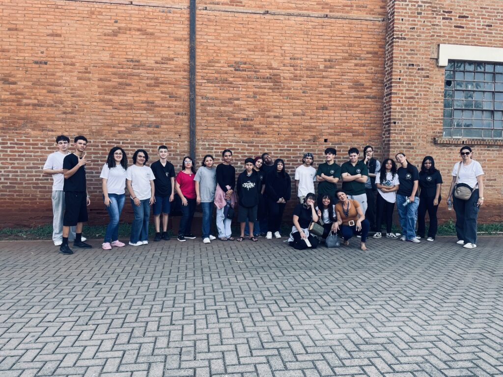 A foto mostra um grupo grande de pessoas posando em frente a uma construção de tijolos aparentes. A maioria está em pé, enquanto algumas pessoas estão agachadas na parte da frente. O grupo é formado principalmente por jovens, acompanhados por alguns adultos, e muitos usam camisetas pretas ou brancas com um símbolo estampado. O local tem uma janela grande com vidros pequenos à direita, paredes de tijolos em tons alaranjados e um piso externo de blocos cinza. As pessoas parecem estar em um passeio, atividade escolar ou encontro de grupo.