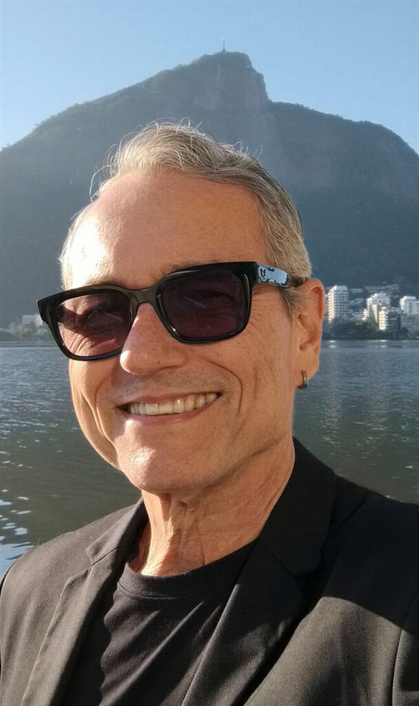 Esta imagem mostra um homem de meia-idade, com cabelos grisalhos curtos e um sorriso simpático, posando para uma selfie. Aqui estão os detalhes: O Homem: Ele usa óculos de sol escuros de armação preta e um pequeno brinco de argola na orelha esquerda. Está vestindo uma camiseta preta básica por baixo de um paletó ou blazer preto, conferindo um visual casual e elegante ao mesmo tempo. O Cenário: O fundo é inconfundível: trata-se da Lagoa Rodrigo de Freitas, no Rio de Janeiro. É possível ver a água da lagoa refletindo a luz do dia e, ao fundo, a silhueta imponente do morro do Corcovado, com o Cristo Redentor visível no topo, além dos prédios típicos da orla carioca à direita. A Atmosfera: A foto tem uma iluminação de fim de tarde, que cria um brilho suave no rosto e destaca o cenário icônico, transmitindo uma sensação de lazer e descontração.