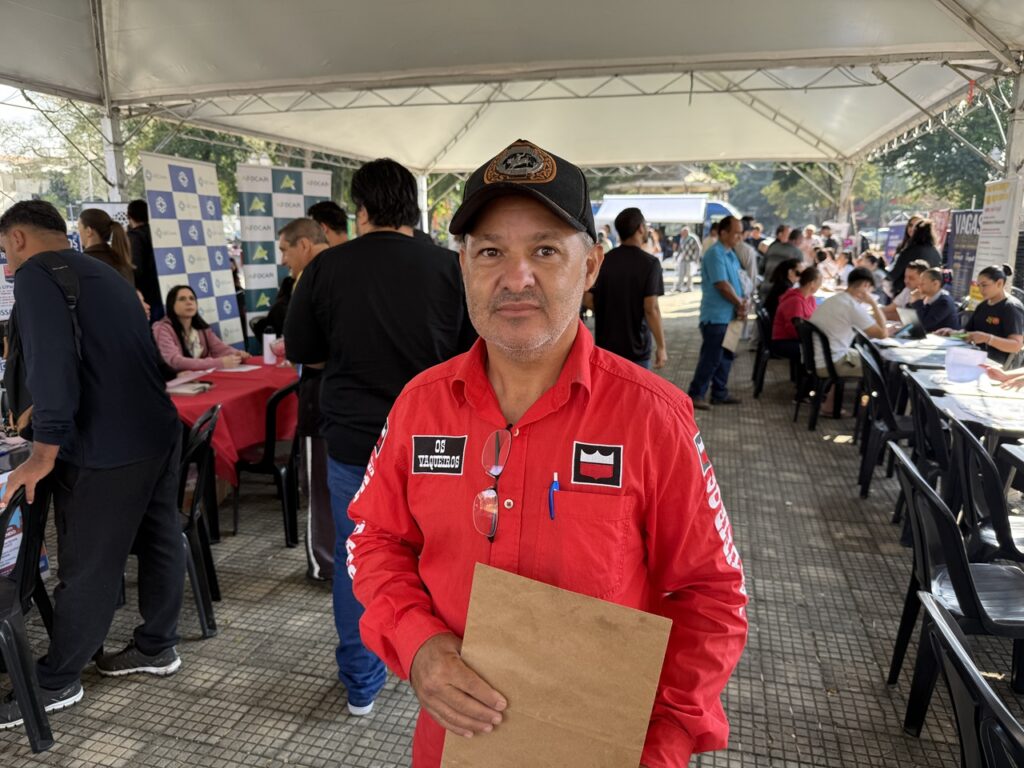 A imagem mostra um homem de meia-idade em pé sob uma tenda branca durante um evento ao ar livre, provavelmente uma feira de empregos ou ação comunitária. Ele veste uma camisa vermelha com bordados e logotipos — incluindo o nome “Os Vaqueiros” — e um boné preto com emblema. Segura uma pasta de papel pardo e olha diretamente para a câmera com expressão serena.