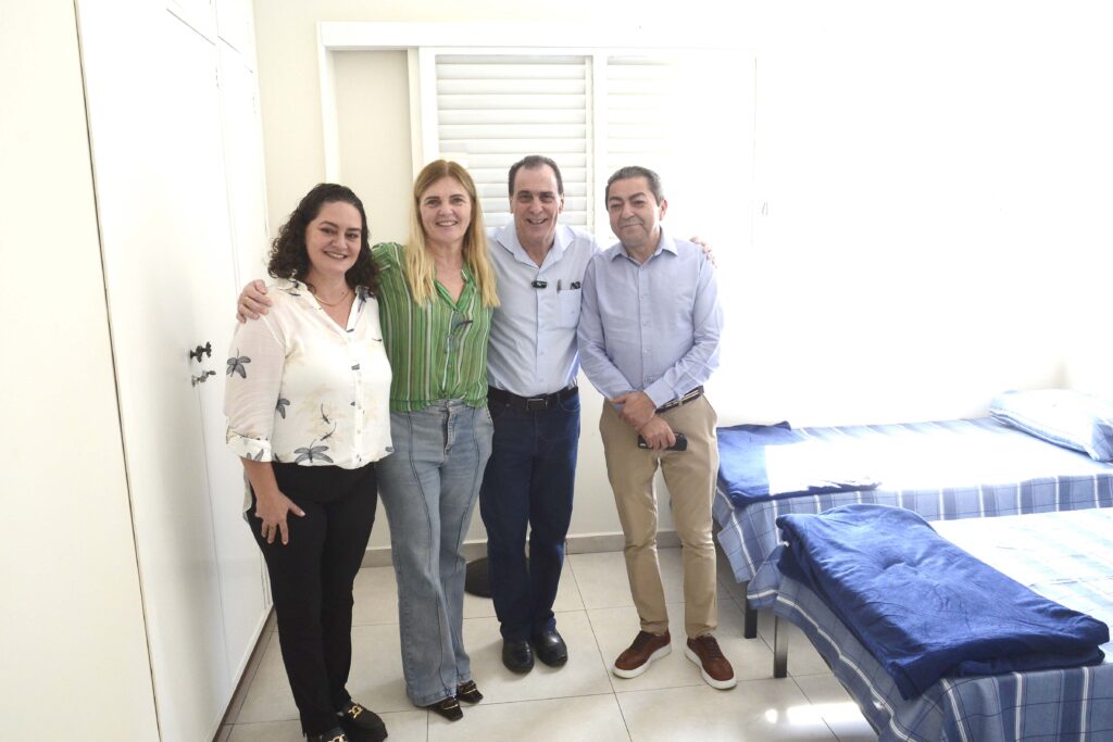 A foto mostra quatro pessoas em uma sala da Residência Inclusiva, posando lado a lado e sorrindo para a câmera. Ao fundo, há duas camas de solteiro organizadas, com roupas de cama em tons de azul, além de uma janela e armários. O ambiente é claro, organizado e bem iluminado.
