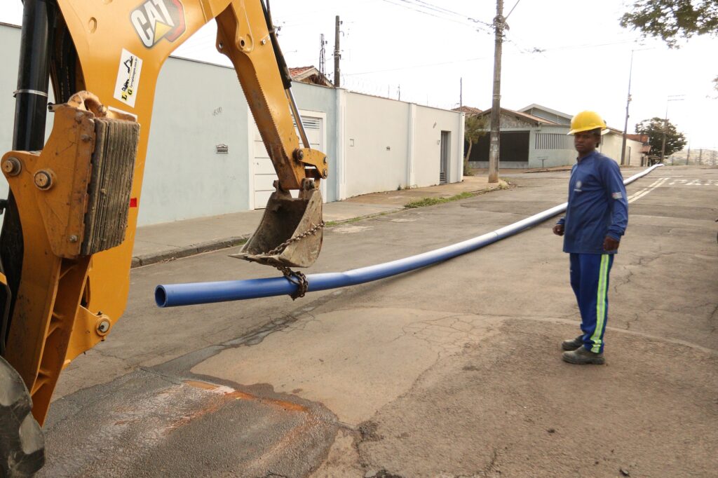 Um trabalhador de capacete amarelo observa enquanto uma escavadeira amarela levanta um longo tubo azul na rua. A imagem mostra uma cena de construção em uma rua residencial com casas ao fundo. No lado esquerdo da imagem, uma grande escavadeira amarela, com o logotipo "CAT" visível, ocupa uma parte significativa do quadro. A lança da escavadeira está erguida, e sua concha está levantando um longo tubo azul que se estende pela rua. A concha está presa ao tubo com uma corrente grossa. O tubo azul é liso e parece ser feito de plástico. Em pé na rua, à direita do tubo, há um trabalhador. Ele usa um capacete de segurança amarelo brilhante, uma blusa azul de manga comprida com um logotipo branco e azul no braço esquerdo, e calças azuis com uma listra verde fluorescente nas laterais. Ele está usando botas de trabalho escuras. O trabalhador está olhando para a escavadeira e o tubo, parecendo supervisionar a operação. Seu corpo está ligeiramente virado para a esquerda, com as mãos em posição neutra. O chão é de asfalto, e há manchas de água ou lama em algumas áreas, sugerindo que o trabalho pode estar relacionado a instalações de água ou drenagem. Ao fundo, há casas com paredes em tons de azul claro e branco. Uma das casas tem um portão de metal escuro e uma cerca de arame farpado no topo do muro. O céu está claro, quase branco, indicando um dia nublado ou um ambiente com luz difusa. A atmosfera geral da imagem é de trabalho em andamento, com um senso de atividade e propósito profissional. O humor é prático e direto, focado na tarefa em questão.