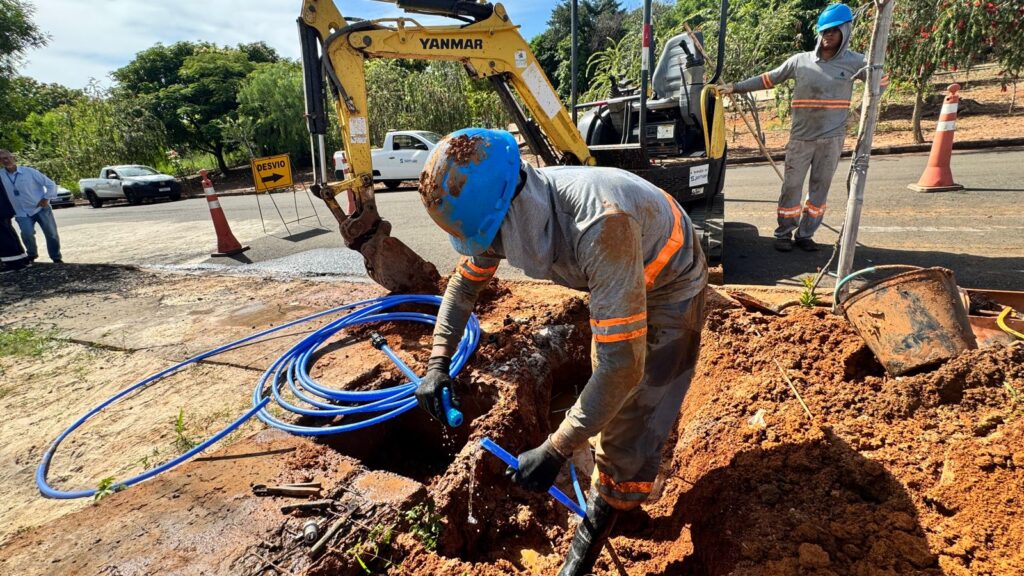 A construção ou reparo de uma tubulação está ocorrendo ao ar livre, com terra vermelha exposta. No centro da imagem, um trabalhador está curvado, vestindo um capacete azul e uniforme cinza com listras laranja reflexivas nas mangas e nas pernas. Ele está trabalhando em uma vala, segurando uma mangueira azul. Salpicos de lama cobrem parcialmente seu capacete e uniforme. Uma escavadeira amarela com a marca Yanmar está posicionada atrás dele, parcialmente visível na cena. No lado direito, outro trabalhador, também de capacete azul e uniforme semelhante, está em pé, apoiado em um poste. No primeiro plano e no lado esquerdo, há uma grande quantidade de mangueira azul enrolada no chão. Ferramentas manuais como alicates e um balde sujo e enferrujado estão espalhados perto da área de trabalho. O fundo da imagem mostra uma estrada pavimentada, com um cone de trânsito laranja e um sinal de "DESVIO" com uma seta apontando para a direita. Uma picape branca e outros veículos estão visíveis ao longe, juntamente com árvores verdes exuberantes e um céu parcialmente nublado. A composição foca nos trabalhadores e no equipamento, destacando a natureza do trabalho em andamento. As cores predominantes são o marrom da terra, o amarelo da escavadeira, o azul das mangueiras e capacetes, e o cinza e laranja dos uniformes. A iluminação sugere um dia claro e ensolarado.