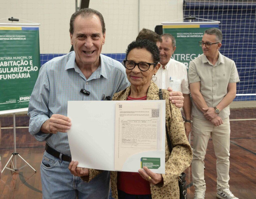 A imagem mostra um momento de entrega de documento oficial em um evento da Secretaria Municipal de Habitação e Regularização Fundiária de Piracicaba. Em primeiro plano, um homem e uma mulher sorriem enquanto exibem um certificado de regularização fundiária, simbolizando a conquista da propriedade legal de um imóvel. Ao fundo, outras pessoas participam da cerimônia, reforçando o caráter público e comunitário da ação. O ambiente é um ginásio, com banners institucionais e clima de celebração.