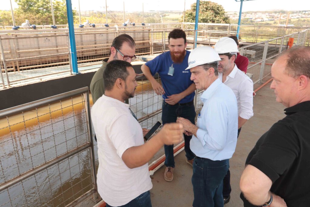 A imagem mostra um grupo de pessoas reunido em uma passarela elevada, aparentemente durante uma visita técnica a uma estação de tratamento de água ou de efluentes. Algumas pessoas usam camisetas brancas com logotipo e crachás, enquanto outras vestem camisetas azuis. Ao fundo, há grandes tanques retangulares com água, passarelas metálicas, grades de proteção, tubulações e estruturas industriais. O grupo parece observar ou receber explicações sobre o funcionamento das instalações. A foto está na orientação vertical e apresenta certo desfoque de movimento.