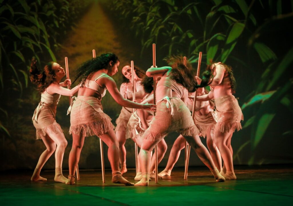 A imagem mostra um grupo de dançarinas se apresentando em um palco. Elas usam figurinos em tons de bege, com saias de franjas e meias, e seguram longos bastões de madeira durante a coreografia. Ao fundo, há um cenário que lembra uma plantação ou floresta, iluminado em tons verdes e alaranjados. As bailarinas estão em diferentes posições, formando uma composição dinâmica e teatral.