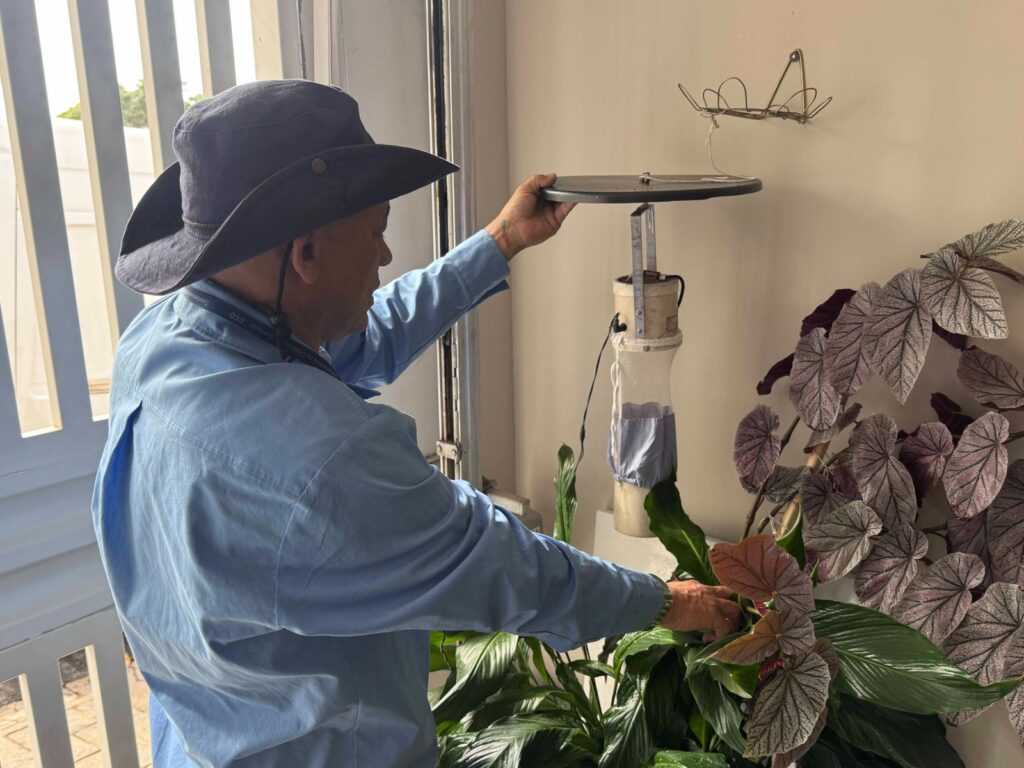 Um homem de chapéu ajusta cuidadosamente um dispositivo cilíndrico suspenso perto de uma planta ornamental, em um ambiente interno que parece ser uma área de serviço ou varanda iluminada. A composição da cena é focada no lado direito, onde o homem interage diretamente com o objeto suspenso e a folhagem. Ele está posicionado de costas e de lado para a câmera, criando uma diagonal visual que conecta suas mãos ao dispositivo e à planta. O enquadramento é em plano médio, permitindo observar tanto a ação técnica do ajuste do equipamento quanto a proximidade com a vegetação. A verticalidade do dispositivo e a disposição das folhas criam um equilíbrio entre os elementos técnicos e naturais dentro do espaço restrito. O protagonista é um homem de pele morena, vestindo uma camisa de botão azul claro de manga comprida e um chapéu de sol de abas largas na cor azul marinho, com uma corda de ajuste sob o queixo. Suas mãos estão ativas: a mão esquerda segura a parte superior de um disco preto que serve de tampa ou suporte para o dispositivo, enquanto a mão direita toca suavemente as folhas de uma planta próxima. Sua postura é de concentração, com o corpo inclinado em direção ao objeto, demonstrando um cuidado meticuloso na manipulação do equipamento. O dispositivo em questão parece ser uma armadilha ou um coletor de insetos feito de um tubo cilíndrico branco com uma tela de malha acoplada, possivelmente utilizado para monitoramento ambiental ou botânico. Ao lado, encontra-se uma begônia com folhas grandes e texturizadas em tons de cinza-esverdeado e bordas arroxeadas, exibindo um contraste vívido com o verde escuro de uma planta do tipo lírio-da-paz logo abaixo. A execução técnica da imagem reflete uma captura fotográfica cotidiana, com foco nítido nos detalhes táteis das plantas e na estrutura do objeto. A cena ocorre em um ambiente fechado, iluminado pela luz natural que entra por uma porta ou janela com grades brancas à esquerda. A iluminação é suave e difusa, criando um clima de trabalho doméstico ou manutenção técnica. O fundo é neutro, com uma parede clara que destaca as cores das plantas. A atmosfera é de tranquilidade e atenção aos detalhes, transmitindo uma sensação de cotidiano produtivo onde o homem se dedica à verificação de seus elementos de cultivo, unindo a jardinagem a um componente de observação científica ou prática.