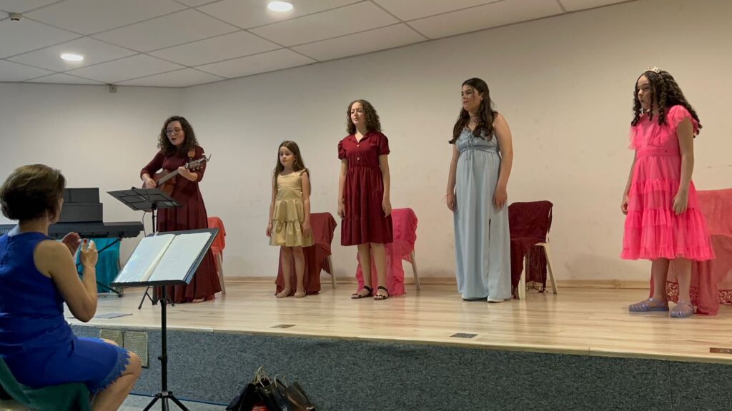 Uma pequena apresentação musical ocorre em um palco, onde uma mulher toca violão enquanto quatro meninas estão alinhadas e prontas para cantar, sob a supervisão de uma instrutora sentada à frente. A composição da cena é organizada de forma linear, com as quatro meninas dispostas horizontalmente no centro do palco, criando uma narrativa visual de uma performance em grupo. À esquerda, a musicista está posicionada com seu violão e partitura, estabelecendo o ritmo, enquanto, em primeiro plano e fora do palco, uma instrutora de azul observa atentamente, possivelmente regendo ou guiando o grupo. O espaçamento entre as participantes permite uma visão clara de cada indivíduo, enquanto cadeiras estrategicamente colocadas atrás delas adicionam volume e profundidade ao cenário. As quatro meninas apresentam idades variadas e vestimentas distintas: a primeira à esquerda veste um vestido dourado curto; a segunda usa um vestido vermelho midi; a terceira, mais alta, veste um longo vestido azul claro; e a última, à direita, usa um vibrante vestido rosa com babados. Todas mantêm uma postura ereta e séria, típica de quem aguarda o início de uma música. A musicista, à esquerda, usa um vestido longo bordô e está concentrada no instrumento, enquanto a instrutora em primeiro plano, de costas, veste um elegante vestido azul, com sua postura sugerindo uma orientação ativa. Esta é uma fotografia digital que documenta um evento social ou cultural, possivelmente um recital escolar ou de igreja. A execução é direta, focada em registrar o momento com nitidez. Não há uma estilização artística complexa; em vez disso, a imagem prioriza a clareza dos sujeitos e do ambiente, utilizando uma iluminação interna e uniforme vinda do teto, que elimina sombras duras e garante que cada detalhe das expressões e das roupas seja visível e bem iluminado. O cenário é um ambiente fechado com paredes claras e um piso de madeira que compõe o palco, conferindo uma estética sóbria e funcional. A luz artificial é constante e clara, criando uma atmosfera de ensaio ou apresentação formal. Elementos como os suportes de partitura e o teclado ao fundo reforçam o contexto musical. O clima geral é de seriedade e expectativa, capturando um instante de disciplina artística e preparação, onde a simplicidade do ambiente destaca a presença e o papel de cada participante no grupo.