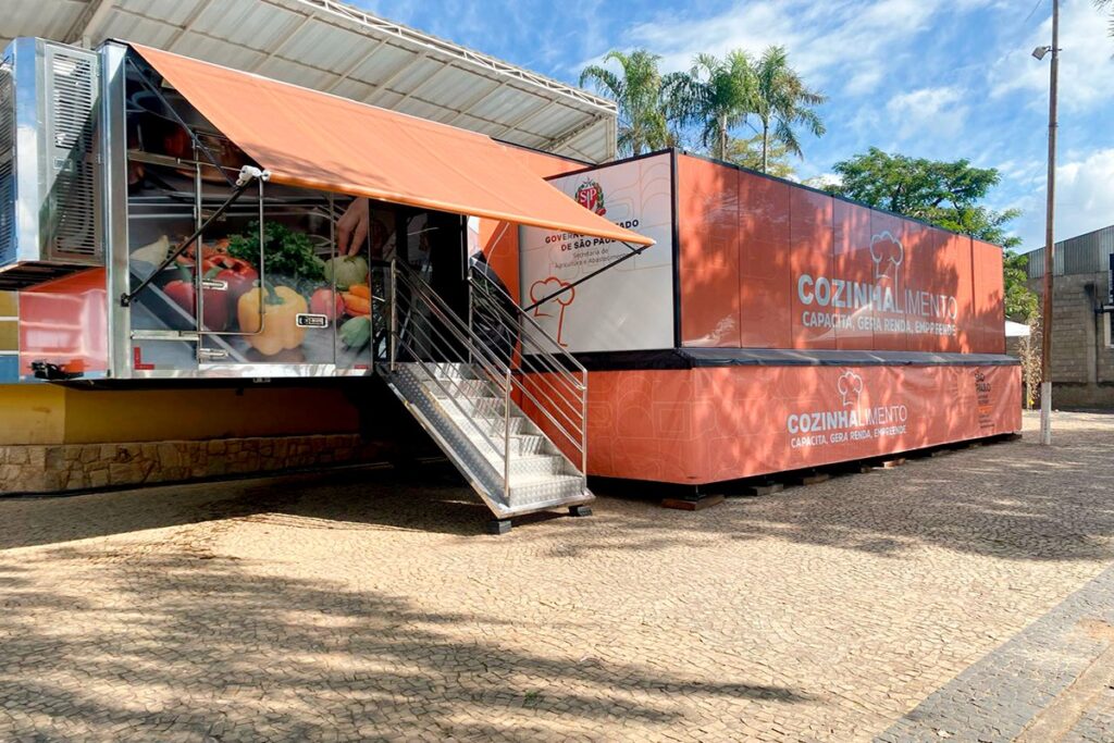 A imagem mostra uma unidade móvel de capacitação culinária, instalada em um caminhão e um contêiner laranja. Na lateral, lê-se “Cozinhalimento – Capacita, gera renda, empreende”. Também aparecem referências ao Governo do Estado de São Paulo e à Secretaria de Agricultura e Abastecimento. O caminhão tem uma cozinha equipada, com imagens de frutas e verduras, uma cobertura retrátil laranja e uma escada metálica de acesso. A estrutura está estacionada em uma área externa pavimentada, sob um céu ensolarado e cercada por árvores.